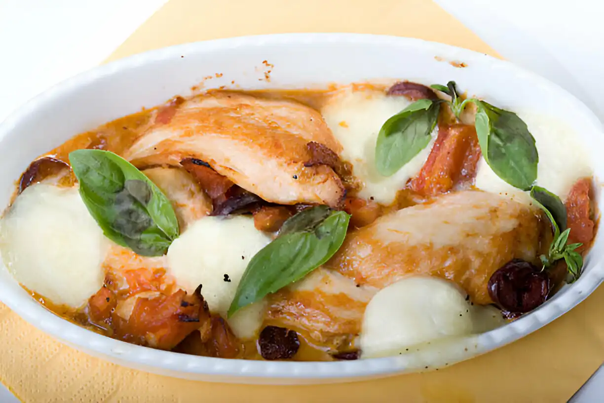 A delicious baked dish featuring chicken pieces, mozzarella cheese, and fresh basil leaves, served in a white ceramic dish.